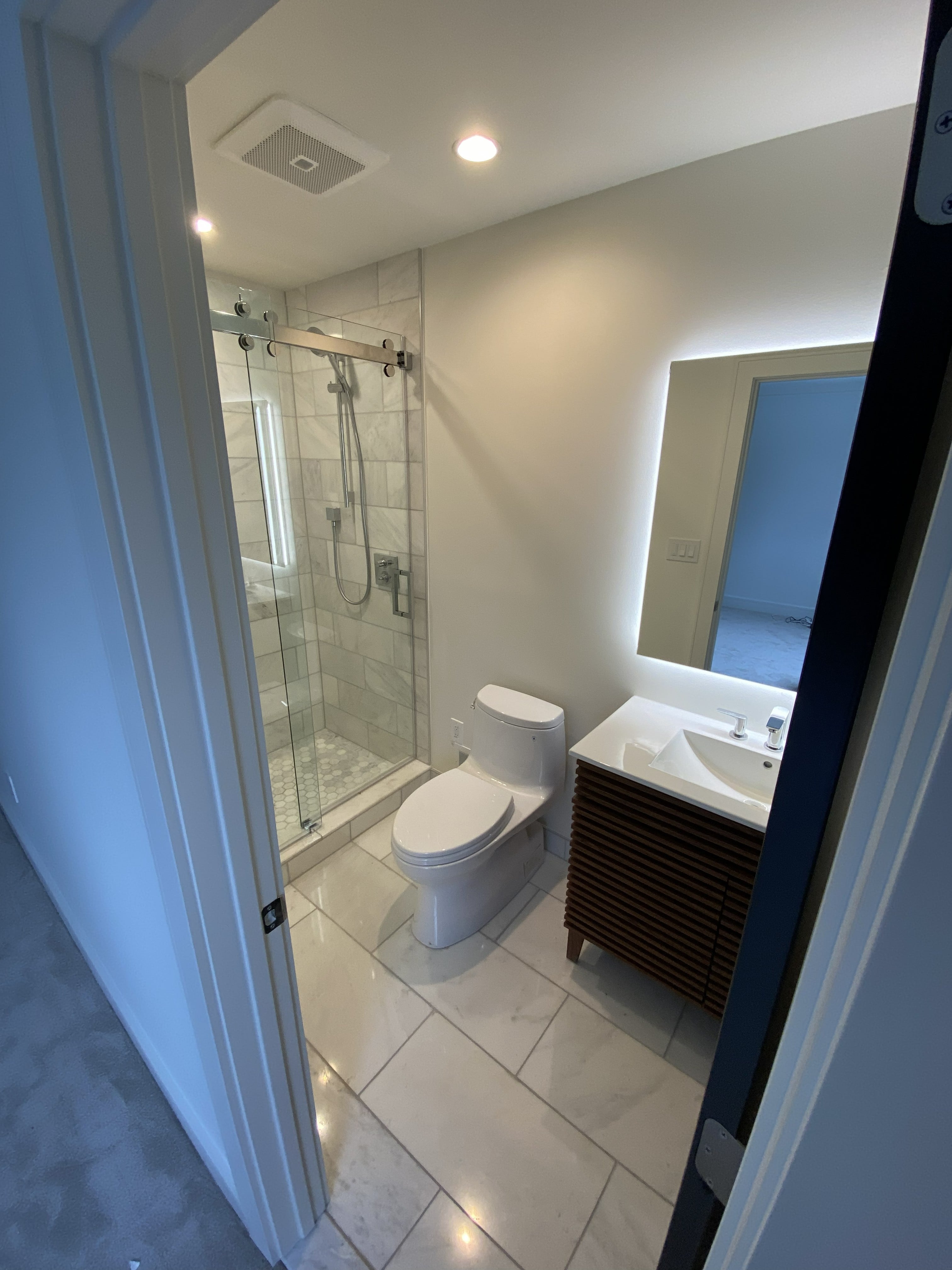 Completed bathroom renovation with frameless sliding glass shower, marble tile surround, hex mosaic floor, walnut slatted vanity, and backlit LED mirror by JD Contracting LLC