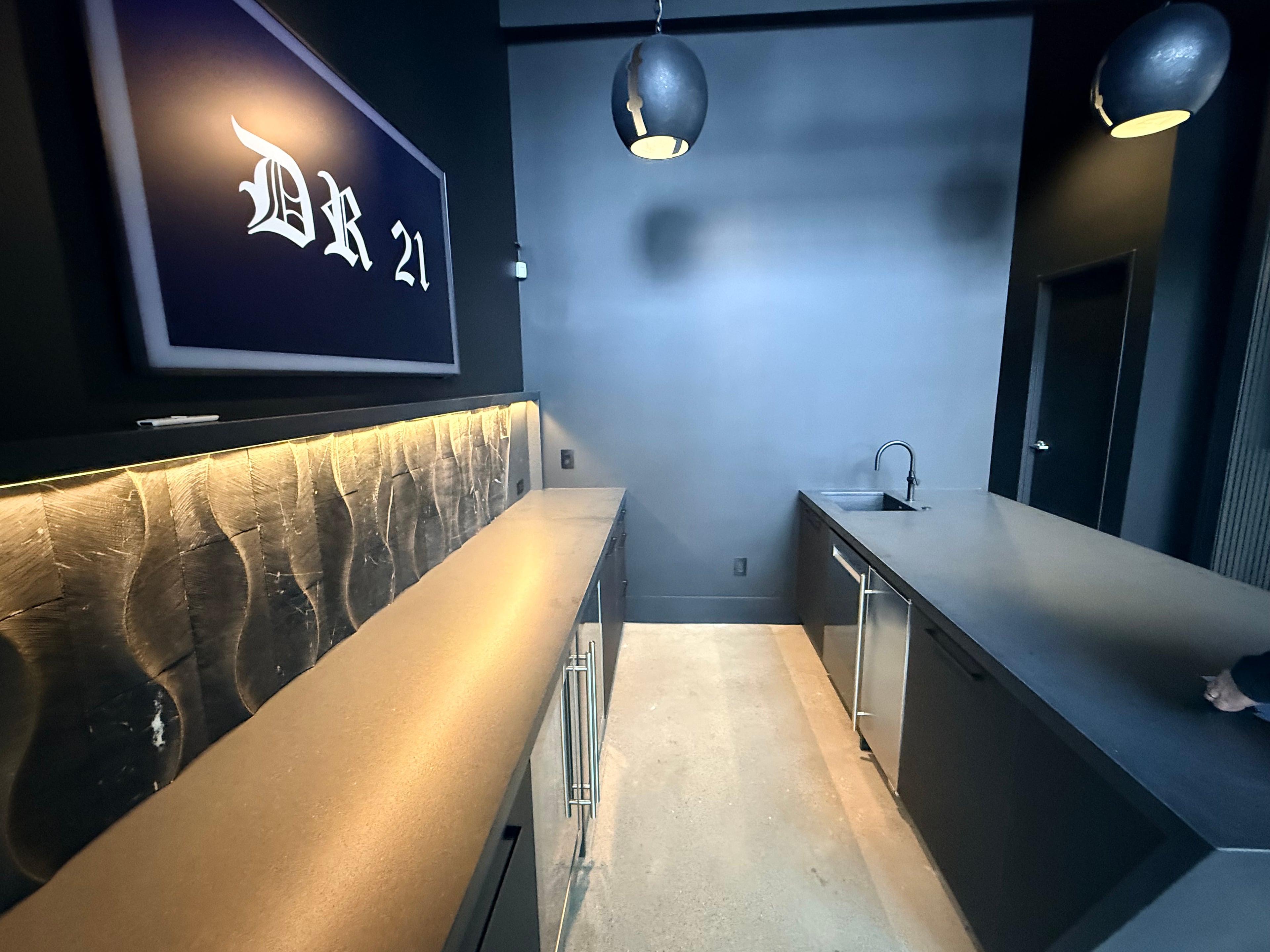 DR 21 commercial bar and kitchenette build-out showing textured gold metallic backsplash with LED underlighting, concrete countertops, matte black cabinetry with bar pulls, undermount sink with matte black faucet, framed DR 21 signage, and globe pendant lights: commercial build-out by JD Contracting LLC