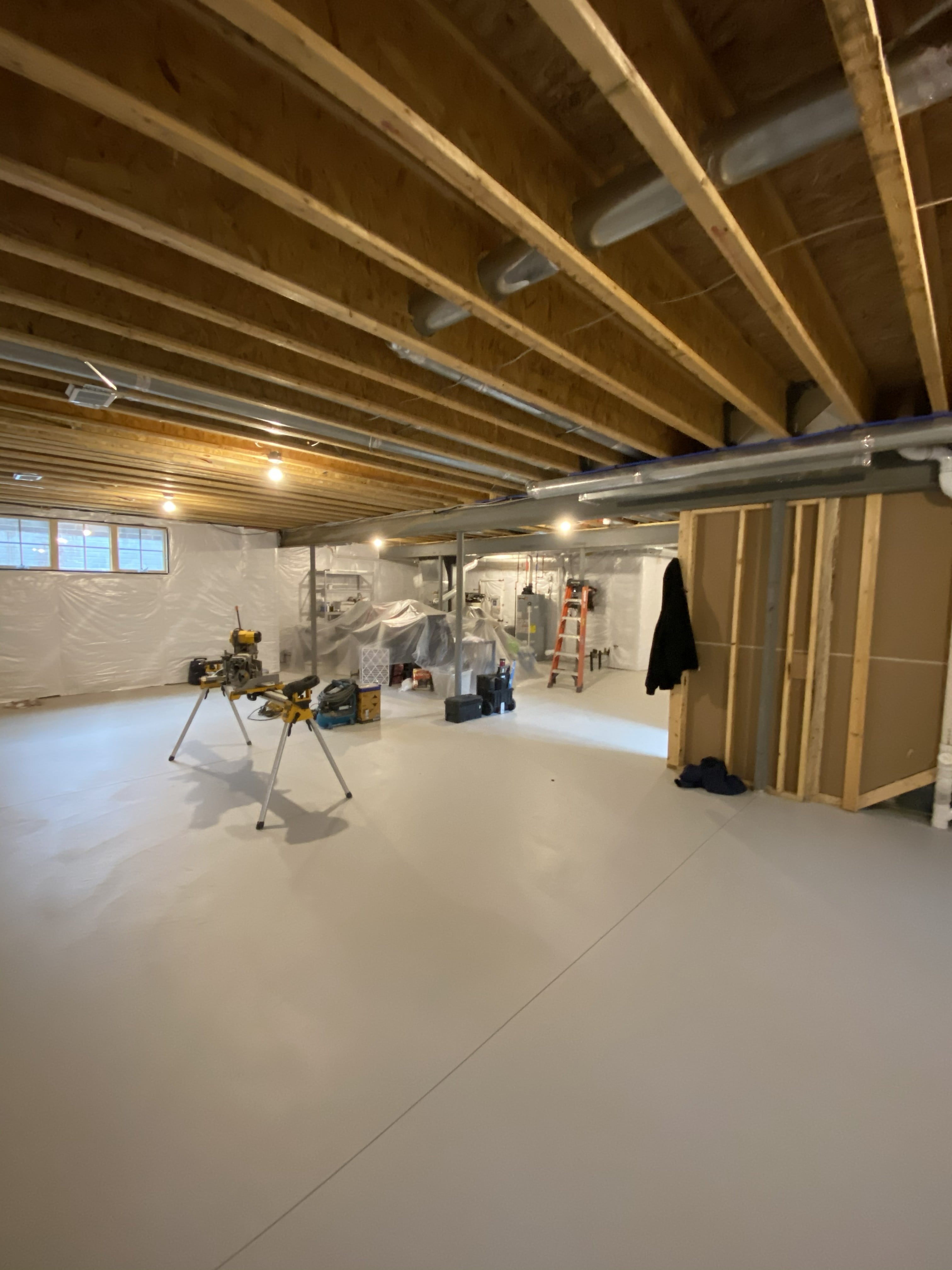 Basement conversion in progress showing painted concrete floor, exposed wood floor joists, new framed partition wall, miter saw on stand, vapor barrier on foundation walls, egress window, and HVAC rough-in ductwork: by JD Contracting LLC