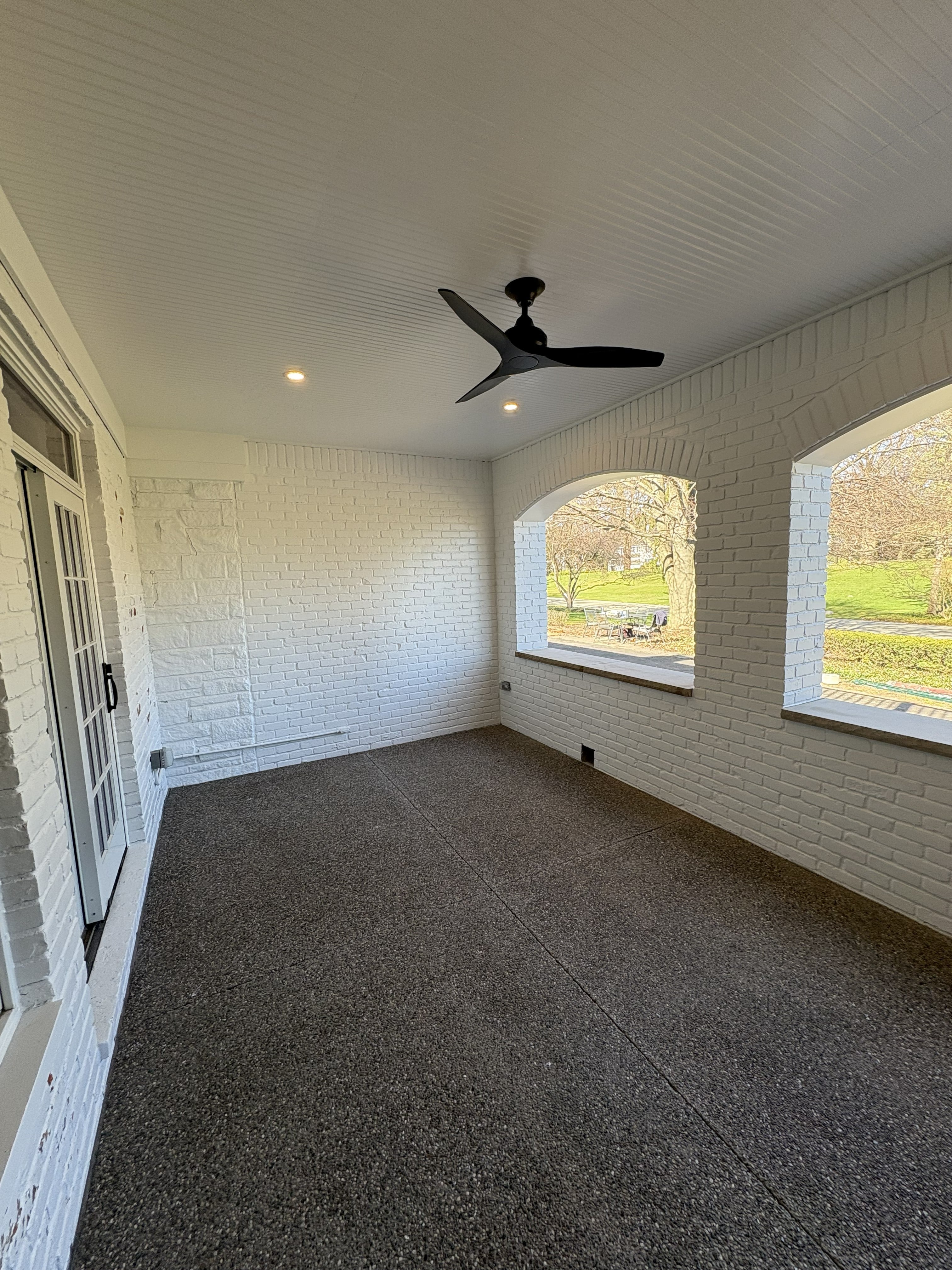 Completed enclosed room addition with white painted brick walls, white beadboard ceiling with recessed lighting and ceiling fan, two large arched picture windows overlooking a landscaped yard, and dark aggregate concrete floor: built by JD Contracting LLC