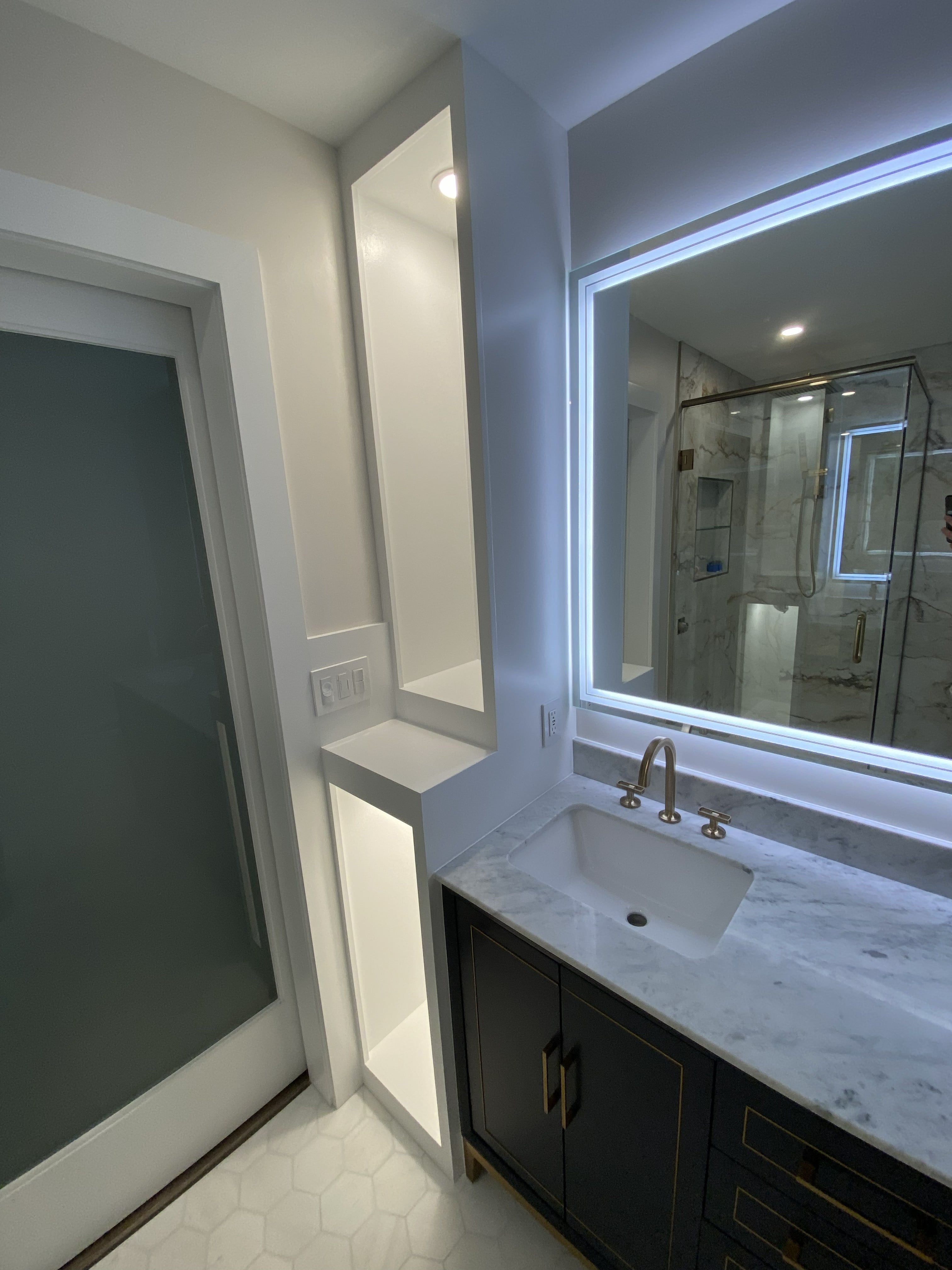 Primary bathroom remodel with dark navy vanity cabinet and gold hardware, Carrara marble countertop, brushed gold widespread faucet, large LED backlit mirror, walk-in glass shower with marble tile walls, and white hexagonal floor tile: by JD Contracting LLC