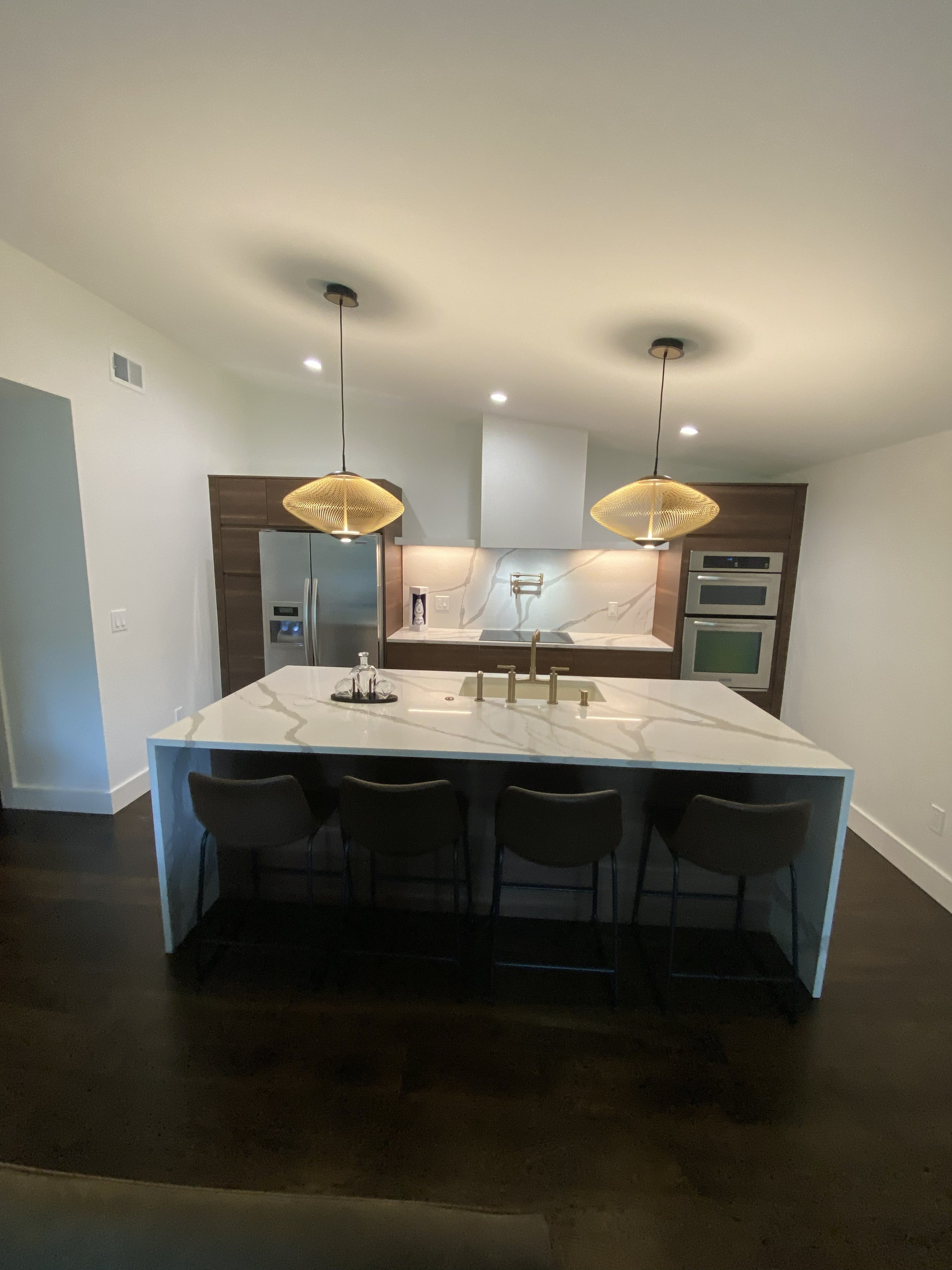 Open-concept kitchen remodel with large waterfall quartz island, four dark upholstered bar stools, brushed gold island faucet, two woven saucer pendant lights, walnut flat-panel upper cabinets flanking a white range hood, stainless french door refrigerator, double wall ovens, and dark hardwood floors: by JD Contracting LLC